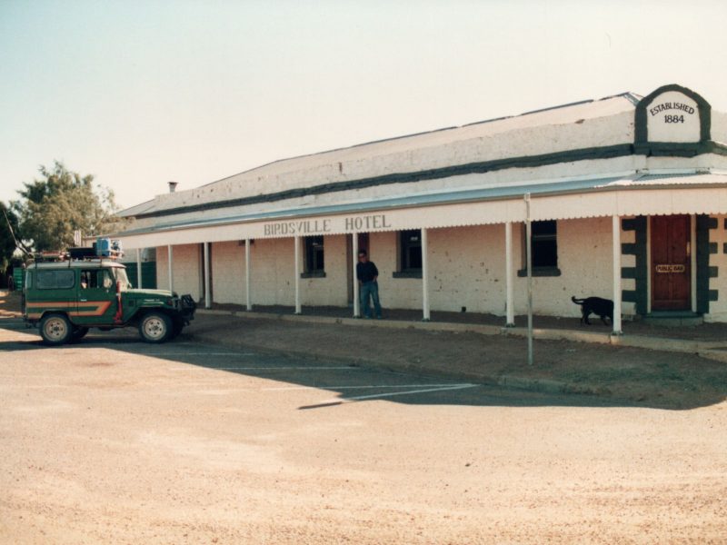 The Birdsville Hotel in 1987. A man stands in front of the Hotel, leaning against a beam. It's a bright, sunny day, a dog is walking around nearby and the sign on the door reads, "Public Bar".
