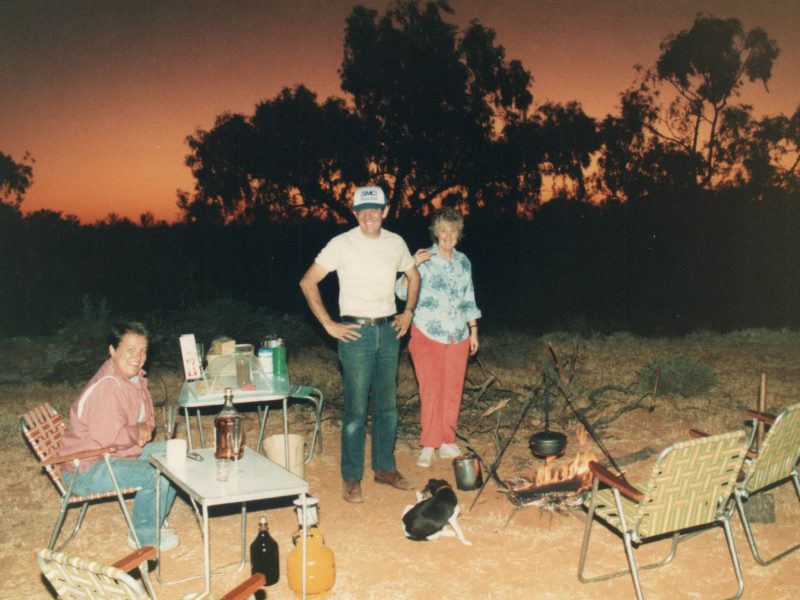 Three adults are relaxing in front of a campfire in the outback. They are happy and sharing a drink, including billy tea and beer.