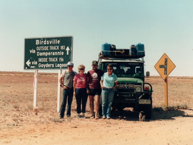 Two couples stand in front of a green landrover 4WD in a dusty landscape. Beside them is a road sign pointing to Birdsville.